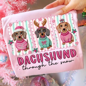 May include: A pink t-shirt with a design of three dachshunds dressed for Christmas. Each dog wears a sweater and festive accessories like a Santa hat, antlers, and a gift. The text "DACHSHUND through the snow" is in pink.