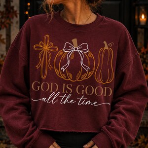 Puede incluir: Una sudadera de manga larga color granate con un estampado gr&aacute;fico de una cruz, una calabaza con un lazo blanco y una calabaza alargada. El texto "GOD IS GOOD" aparece en letras marrones sobre el texto en cursiva blanca "all the time".