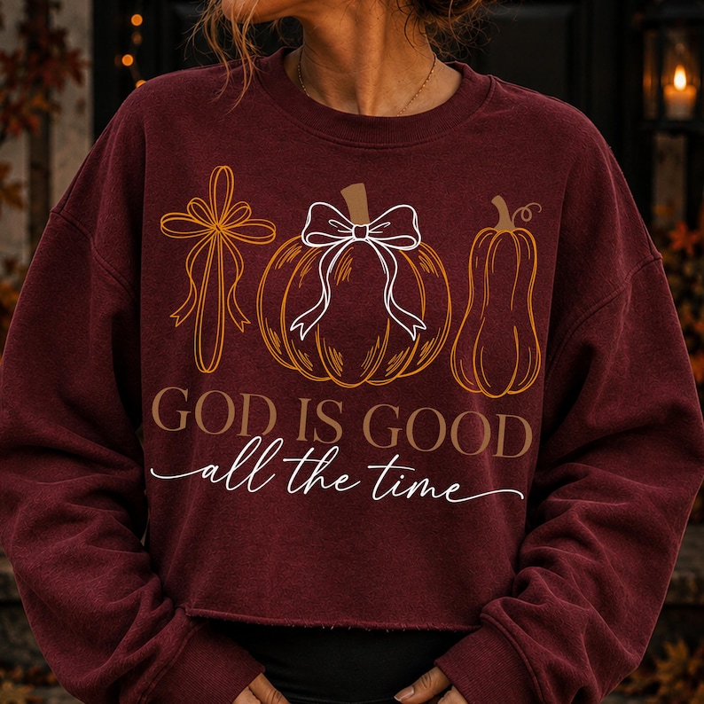 Puede incluir: Una sudadera de manga larga color granate con un estampado gr&aacute;fico de una cruz, una calabaza con un lazo blanco y una calabaza alargada. El texto "GOD IS GOOD" aparece en letras marrones sobre el texto en cursiva blanca "all the time".