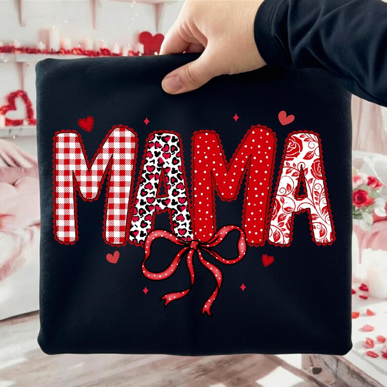 Może przedstawiać: Czarna bluza z napisem "MAMA" w kolorze czerwono-białym. Litera "M" jest w czerwoną i białą kratę, "A" ma wz&oacute;r serca, druga "M" jest czerwona w białe kropki, a ostatnia "A" ma kwiatowy wz&oacute;r. Na dole czerwona kokarda.