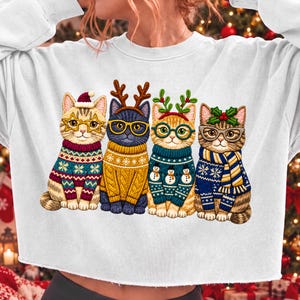 May include: A white long-sleeve sweatshirt featuring a graphic of four cats wearing holiday sweaters and accessories. The cats wear star-shaped glasses, reindeer antlers, and holly, with patterns including snowflakes and snowmen on their sweaters.