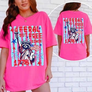 May include: A bright pink t-shirt with a graphic featuring a raccoon dressed as the Statue of Liberty, holding a torch and a tablet, with the text "FERAL BUT FREE SINCE 1776". The design includes blue stripes, stars, and fireworks.