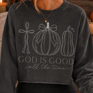 K&ouml;nnte beinhalten: Ein anthrazitfarbenes Langarm-Sweatshirt mit einer wei&szlig;en Strichgrafik, die ein Kreuz, einen K&uuml;rbis mit Schleife und einen Flaschenk&uuml;rbis zeigt. Der Text "GOD IS GOOD all the time" ist darunter in einer Kombination aus Block- und Schreibschrift gedruckt.