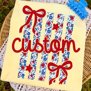 Puede incluir: Una camiseta amarilla clara con la palabra "custom" en rojo. La camiseta presenta rayas verticales con un estampado floral en rojo, blanco y azul, junto con lazos y estrellas rojas. Una pinza para el pelo azul con detalles florales est&aacute; en la esquina superior derecha.