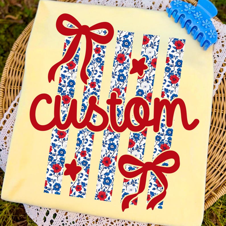 Puede incluir: Una camiseta amarilla clara con la palabra "custom" en rojo. La camiseta presenta rayas verticales con un estampado floral en rojo, blanco y azul, junto con lazos y estrellas rojas. Una pinza para el pelo azul con detalles florales est&aacute; en la esquina superior derecha.