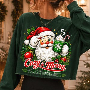 K&ouml;nnte beinhalten: Ein dunkelgr&uuml;nes, bauchfreies Langarm-Sweatshirt mit einer Grafik vom zwinkernden Weihnachtsmann, der einen gr&uuml;nen Becher Kakao mit Sahne und Zuckerstange h&auml;lt. Der Text lautet Cozy & Merry SANTA'S COCOA CLUB mit roten Weihnachtskugeln.