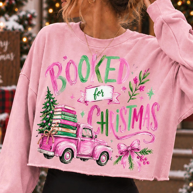 May include: A light pink long-sleeve cropped sweatshirt featuring a graphic print with the text BOOKED for CHRISTMAS. The design includes a pink pickup truck carrying a stack of books and a Christmas tree, accented with a pink bow and greenery.
