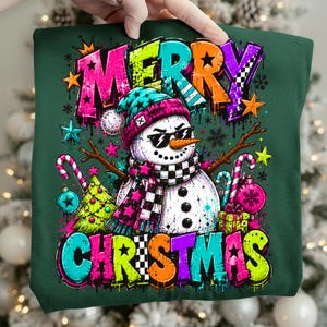Puede incluir: Una sudadera verde oscuro con un gr&aacute;fico de un mu&ntilde;eco de nieve con gafas de sol, un gorro azul verdoso y una bufanda a cuadros blancos y negros. El texto MERRY CHRISTMAS est&aacute; impreso en letras grandes y coloridas de estilo grafiti.
