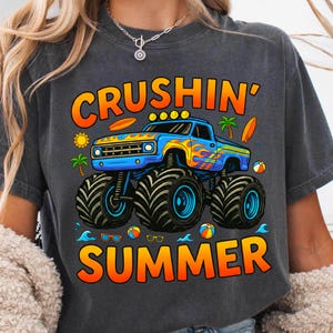 May include: Dark gray t-shirt featuring a blue monster truck with orange and yellow flames, the words "CRUSHIN' SUMMER" in orange and yellow, and summer-themed graphics like surfboards, palm trees, and beach balls.