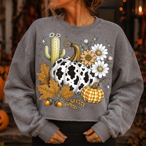 Puede incluir: Una sudadera gris carb&oacute;n de cuello redondo con un estampado gr&aacute;fico de una calabaza con manchas de vaca, una calabaza a cuadros amarillos, un cactus verde, hojas de oto&ntilde;o naranjas, bellotas y margaritas blancas y amarillas con estrellas.