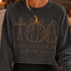 K&ouml;nnte beinhalten: Ein anthrazitfarbenes bauchfreies Sweatshirt mit einer goldenen Linienzeichnung, die ein Kreuz mit Schleife, einen K&uuml;rbis mit Schleife und einen Flaschenk&uuml;rbis zeigt. Der Text "GOD IS GOOD all the time" ist in goldenen Block- und Schreibschriftbuchstaben.