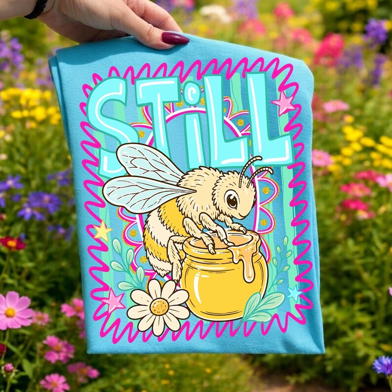 Op de afbeelding: Lichtblauw T-shirt met een cartoon bijenontwerp en het woord "STILL". De bij zit naast een honingpot. Het ontwerp is omlijst door een roze, golvende rand met sterren en bloemen.