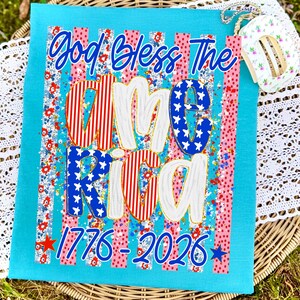 Puede incluir: Un cuadrado de tela turquesa con las palabras "God Bless The America" en escritura azul. La palabra "America" est&aacute; en rojo, blanco y azul con un estampado floral. Los n&uacute;meros "1776-2026" est&aacute;n en la parte inferior.