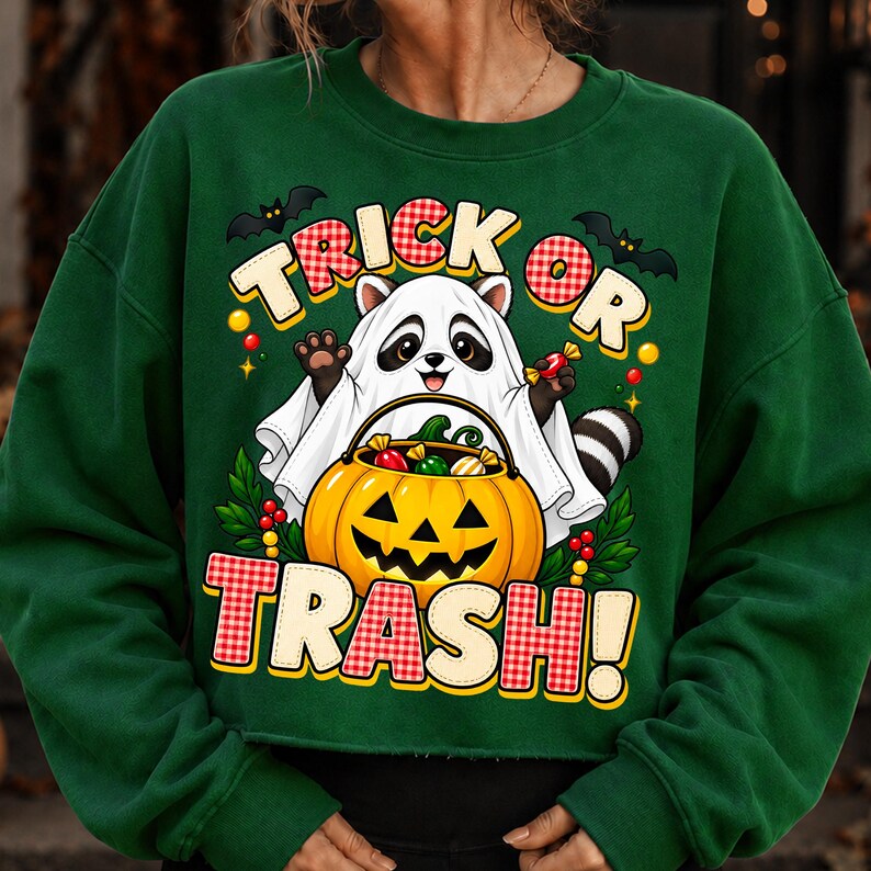 May include: A dark green long-sleeve sweatshirt featuring a graphic of a raccoon wearing a white ghost costume. The raccoon holds a jack-o'-lantern bucket filled with sweets. The text reads TRICK OR TRASH! with red and white chequered accents.