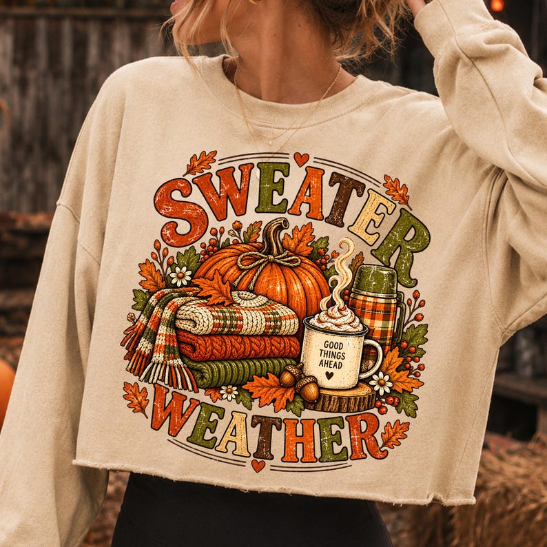 Puede incluir: Una sudadera corta de color beige con mangas largas que presenta un estampado gr&aacute;fico con el texto "SWEATER WEATHER". El dise&ntilde;o incluye una calabaza naranja grande, una pila de su&eacute;teres doblados, una taza con la inscripci&oacute;n "GOOD THINGS AHEAD" y hojas de oto&ntilde;o.