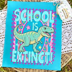 Op de afbeelding: Een turquoise draagtas met de woorden "SCHOOL IS EXTINCT!" in grote, bubbelletters. Een cartoon Tyrannosaurus Rex staat in het midden, omringd door roze sterren en een roze en gele rand. De tas ligt op een geweven oppervlak.