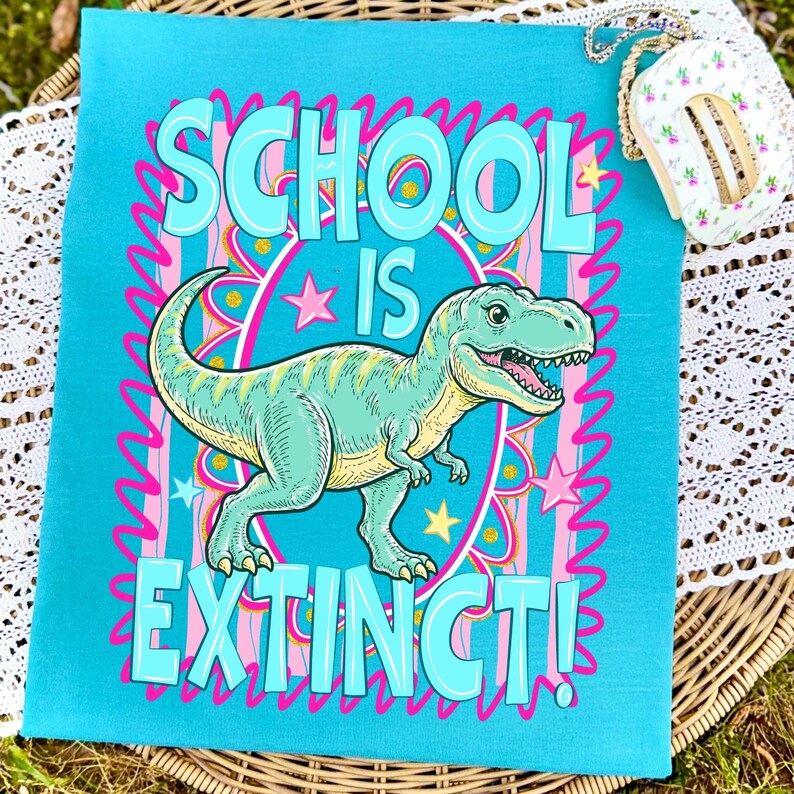 Op de afbeelding: Een turquoise draagtas met de woorden "SCHOOL IS EXTINCT!" in grote, bubbelletters. Een cartoon Tyrannosaurus Rex staat in het midden, omringd door roze sterren en een roze en gele rand. De tas ligt op een geweven oppervlak.