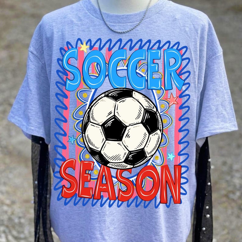 Puede incluir: Camiseta azul claro con las palabras "SOCCER SEASON" en azul y rojo, rodeando un gr&aacute;fico de bal&oacute;n de f&uacute;tbol en blanco y negro. El dise&ntilde;o tiene un estilo ondulado y de dibujos animados con acentos de estrellas.