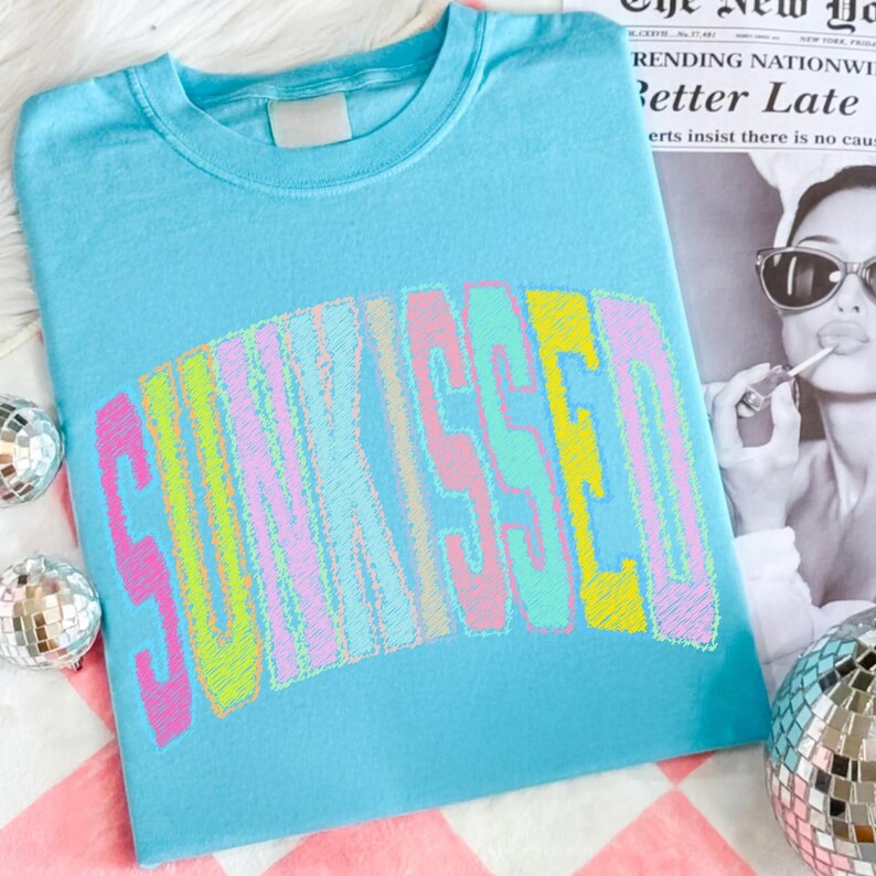 May include: A pale blue t-shirt featuring the word "SUNKISSED" in a multi-coloured, embroidered design. The text is in a curved, arched shape. The shirt is on a pink and white checkered surface.