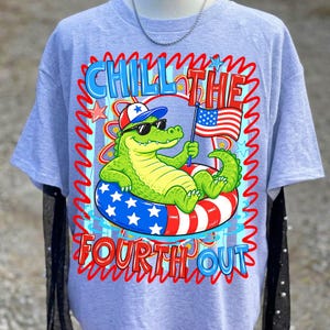 K&ouml;nnte beinhalten: Hellblaues T-Shirt mit einer Grafik eines Cartoon-Alligators mit Sonnenbrille und patriotischem Hut, der in einem rot-wei&szlig;-blauen Schwimmreifen schwimmt. Der Text lautet "Chill the Fourth Out."