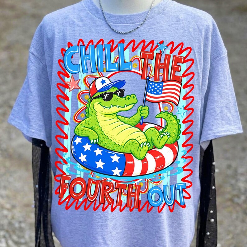 K&ouml;nnte beinhalten: Hellblaues T-Shirt mit einer Grafik eines Cartoon-Alligators mit Sonnenbrille und patriotischem Hut, der in einem rot-wei&szlig;-blauen Schwimmreifen schwimmt. Der Text lautet "Chill the Fourth Out."
