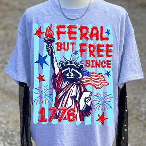May include: A light gray t-shirt featuring a raccoon dressed as the Statue of Liberty, holding a torch and a book, with the text "FERAL BUT FREE SINCE 1776". The design includes red, white, and blue elements, stars, and fireworks.