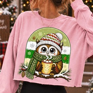 May include: A light pink long-sleeve cropped sweatshirt featuring a graphic of a brown owl wearing a green knit scarf and a red, white, and green striped winter hat. The owl holds a mug of hot cocoa with marshmallows while perched on a pine branch.
