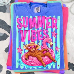 Może przedstawiać: Stos kolorowych koszulek. G&oacute;rna koszulka jest niebieska z r&oacute;żowymi słowami "SUMMER VIBES", a także kresk&oacute;wką szkockiej krowy w okularach przeciwsłonecznych i kapeluszu, unoszącej się na r&oacute;żowym pływaku z flamingiem. Pozostałe koszulki są r&oacute;żowe, pomarańczowe i zielone.