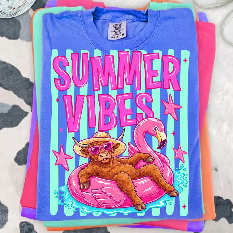 Może przedstawiać: Stos kolorowych koszulek. G&oacute;rna koszulka jest niebieska z r&oacute;żowymi słowami "SUMMER VIBES", a także kresk&oacute;wką szkockiej krowy w okularach przeciwsłonecznych i kapeluszu, unoszącej się na r&oacute;żowym pływaku z flamingiem. Pozostałe koszulki są r&oacute;żowe, pomarańczowe i zielone.
