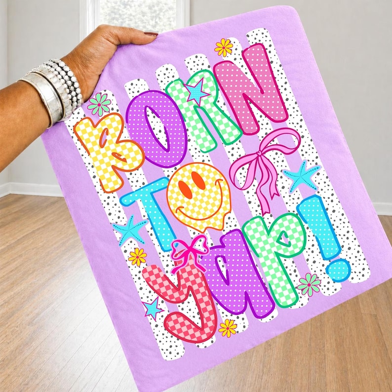 May include: A lilac blanket featuring the phrase "BORN TO YAP!" in vibrant, bubble-style lettering. The design incorporates a smiley face, stars, flowers, and bows. The background has white stripes with black polka dots.