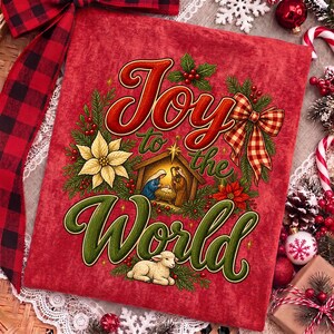 Może przedstawiać: Czerwony kwadratowy kawałek materiału z napisem Joy to the World czerwoną i zieloną kursywą. Wz&oacute;r przedstawia scenę narodzin w stajence, białą gwiazdę betlejemską, owoce ostrokrzewu, gałązki sosny, kokardę w kratę oraz małe białe jagnię.