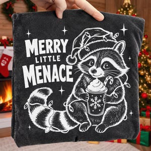 May include: A charcoal gray t-shirt featuring a white graphic of a raccoon wearing a Santa hat and tangled in Christmas lights. The raccoon holds a cup with a snowflake design. The text reads MERRY LITTLE MENACE in bold white lettering.