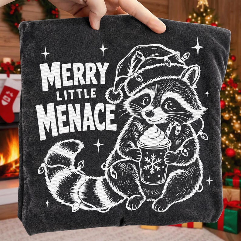 May include: A charcoal gray t-shirt featuring a white graphic of a raccoon wearing a Santa hat and tangled in Christmas lights. The raccoon holds a cup with a snowflake design. The text reads MERRY LITTLE MENACE in bold white lettering.