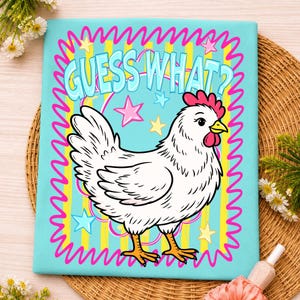 May include: A light blue t-shirt featuring a cartoon chicken with the text "GUESS WHAT?" in light blue. The design includes pink stars and a yellow and pink wavy border.