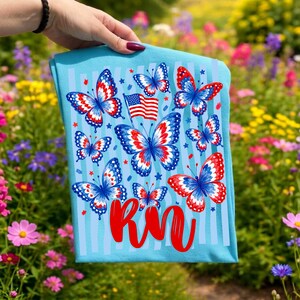 Peut inclure: Un t-shirt bleu clair orn&eacute; de papillons aux ailes rouges, blanches et bleues, d'un petit drapeau am&eacute;ricain et du texte rouge RN. Le motif comprend des rayures verticales bleu clair et de petites &eacute;toiles rouges et bleues.