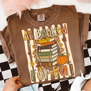 Op de afbeelding: Een bruin t-shirt met korte mouwen en een afbeelding van een stapel boeken en een pompoen. De tekst luidt All Booked For Fall. Het ontwerp bevat herfstachtige elementen zoals bladeren en bloemen tegen een achtergrond van verticale strepen.