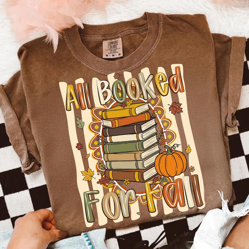 Op de afbeelding: Een bruin t-shirt met korte mouwen en een afbeelding van een stapel boeken en een pompoen. De tekst luidt All Booked For Fall. Het ontwerp bevat herfstachtige elementen zoals bladeren en bloemen tegen een achtergrond van verticale strepen.