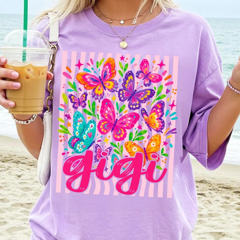 May include: A light purple t-shirt featuring a graphic print of colorful butterflies in shades of pink, orange, yellow, and teal, set against a background of vertical light pink stripes. The word "gigi" is printed in a bold, cursive pink font.