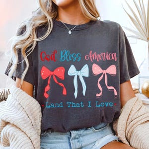 Może przedstawiać: Ciemnoszara koszulka z napisem "God Bless America" w kolorze czerwonym i niebieskim oraz "Land That I Love" w kolorze niebieskim. Koszulka posiada trzy wzory kokardek w kolorze czerwonym, jasnoniebieskim i r&oacute;żowym.
