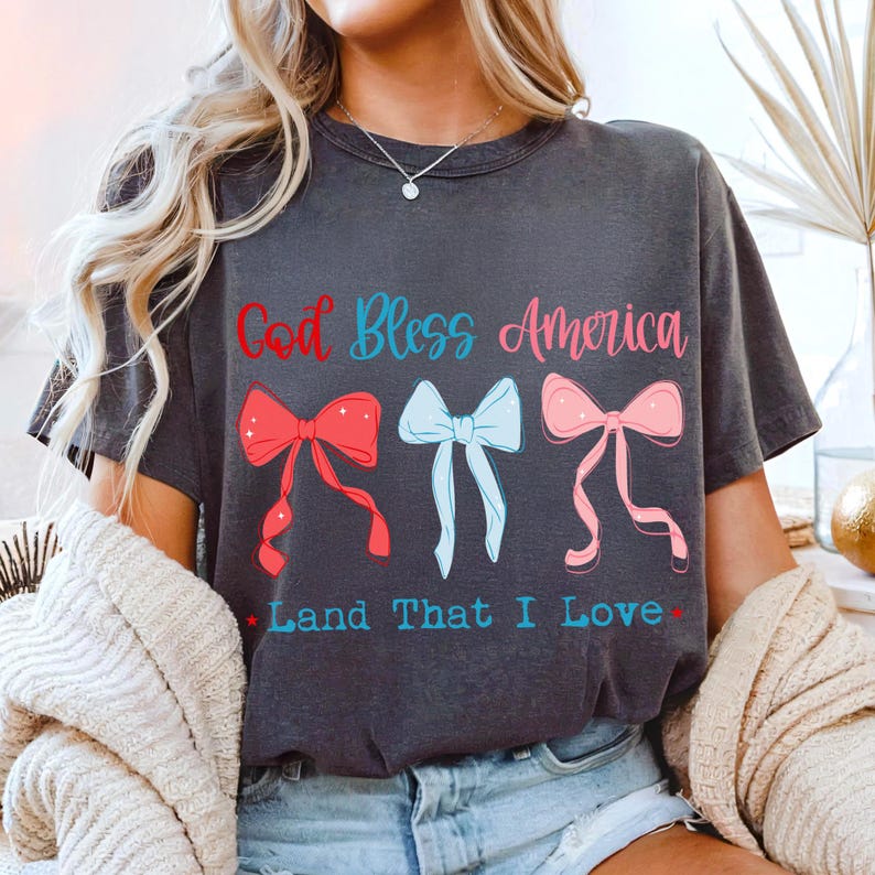 Może przedstawiać: Ciemnoszara koszulka z napisem "God Bless America" w kolorze czerwonym i niebieskim oraz "Land That I Love" w kolorze niebieskim. Koszulka posiada trzy wzory kokardek w kolorze czerwonym, jasnoniebieskim i r&oacute;żowym.