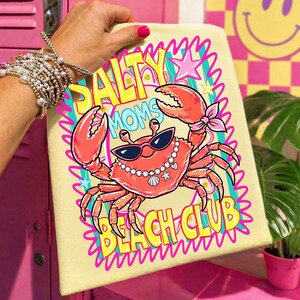 May include: A light yellow beach towel with a cartoon crab wearing sunglasses and a pearl necklace. The crab is surrounded by the words "SALTY MOMS BEACH CLUB" in a colourful, wavy font. Pink and blue accents frame the design.