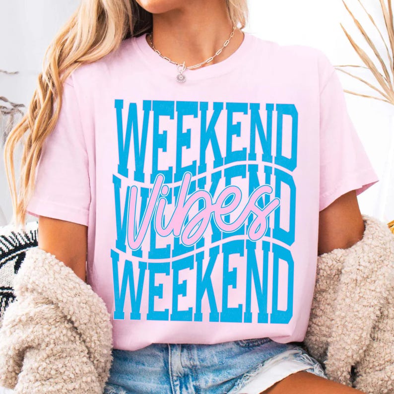 K&ouml;nnte beinhalten: Hellrosa T-Shirt mit dem Schriftzug "WEEKEND Vibes WEEKEND" in Blau und Rosa. Das Shirt hat kurze &Auml;rmel und einen Rundhalsausschnitt. Das Thema ist Freizeitkleidung.