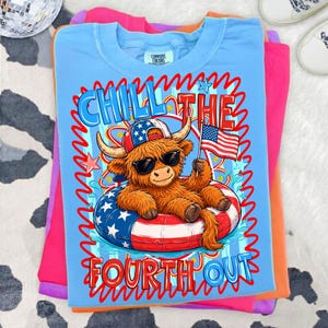 May include: A light blue t-shirt with a graphic of a Highland cow wearing sunglasses and a patriotic hat, floating on an inner tube with an American flag. The text "CHILL THE FOURTH OUT" is displayed in red and blue.