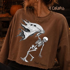 K&ouml;nnte beinhalten: Ein braunes bauchfreies Sweatshirt mit einer schwarz-wei&szlig;en Grafik eines Skeletts, das ein Geisterlaken mit Sternenhimmel-Muster h&auml;lt. Der Text "Sheet Happens..." steht unten und "4 Colors" ist oben zu sehen.