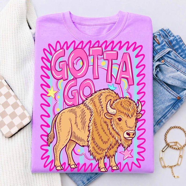 May include: A lilac-colored t-shirt featuring a cartoon bison with the text "GOTTA GO" in pink bubble letters. The bison is tan with brown horns and is surrounded by a pink and blue wavy border with stars.