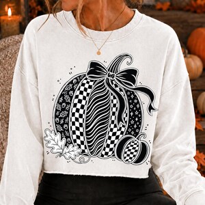 K&ouml;nnte beinhalten: Ein wei&szlig;es Langarm-Sweatshirt mit einer gro&szlig;en schwarzen Grafik eines K&uuml;rbisses, der mit einer Schleife verziert ist. Das K&uuml;rbisdesign umfasst Abschnitte mit Karomustern, Wellenlinien, Eichenbl&auml;ttern, Eicheln und Sternen sowie einen kleineren K&uuml;rbis.