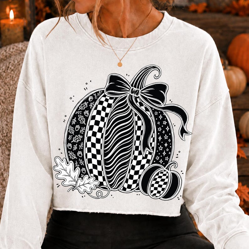 K&ouml;nnte beinhalten: Ein wei&szlig;es Langarm-Sweatshirt mit einer gro&szlig;en schwarzen Grafik eines K&uuml;rbisses, der mit einer Schleife verziert ist. Das K&uuml;rbisdesign umfasst Abschnitte mit Karomustern, Wellenlinien, Eichenbl&auml;ttern, Eicheln und Sternen sowie einen kleineren K&uuml;rbis.
