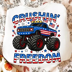 K&ouml;nnte beinhalten: Wei&szlig;es Sweatshirt mit patriotischem Design. Es zeigt einen blauen Monstertruck mit roten und wei&szlig;en Akzenten, amerikanische Flaggen und die Worte "Crushin' Freedom" in Rot, Wei&szlig; und Blau.