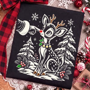 Puede incluir: Una camiseta negra con una ilustraci&oacute;n gr&aacute;fica de un cervatillo con astas marrones y una nariz roja brillante. Un dedo enguantado toca la nariz. El dise&ntilde;o incluye pinos blancos nevados, copos de nieve y un collar con peque&ntilde;as campanas doradas.