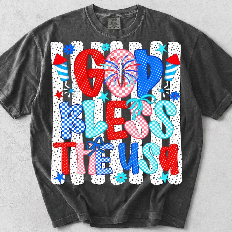 May include: A charcoal grey short-sleeve t-shirt featuring a graphic print with the text GOD BLESS THE USA. The design includes red, white, and blue patterns, polka dots, checkered motifs, fireworks, and stars against a background of vertical stripes.